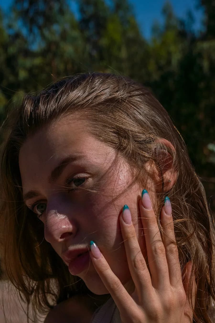 Woman in sunlight with turquoise nails resting on her cheek