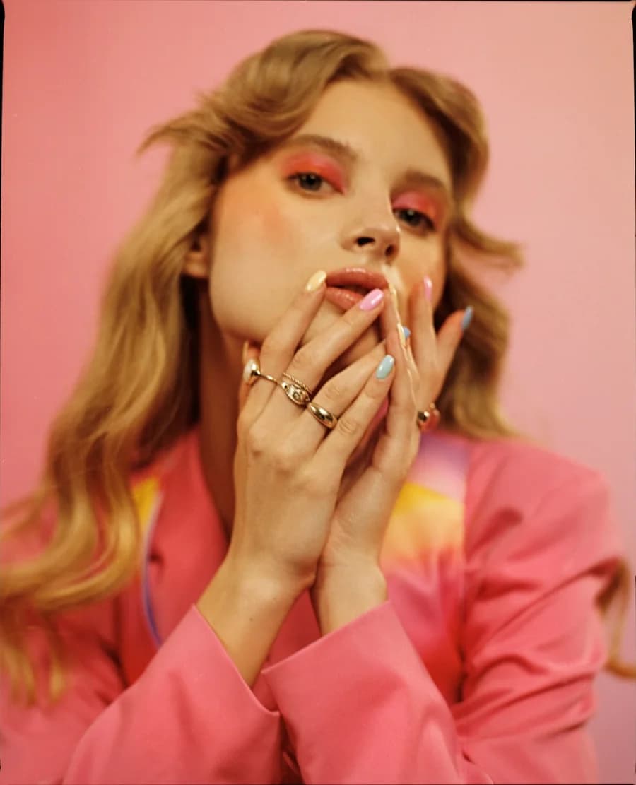 Woman in a pink blazer with pastel nails and gold rings pressed to her lips